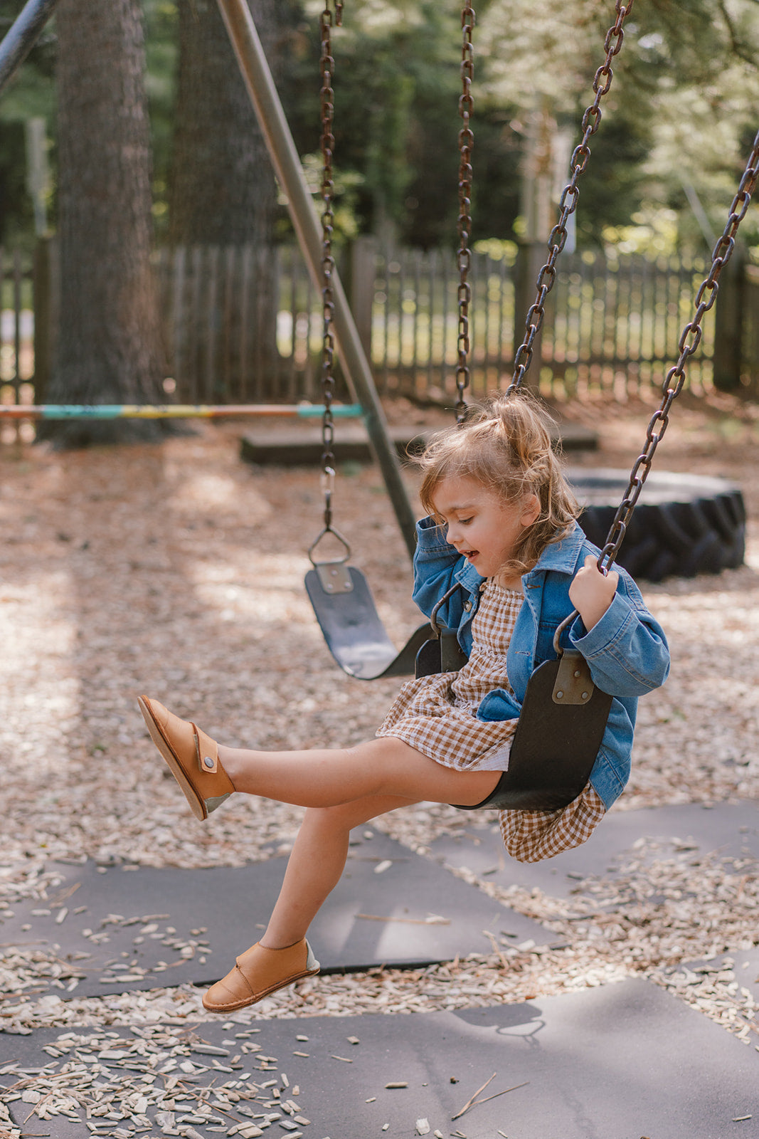 A young girl sitting on a playground swing, wearing a blue denim jacket, a beige and white checkered dress, and tan leather shoes, with trees and a fence in the background. Evergreen Leather shoes by Common Tread shoes featured. 
